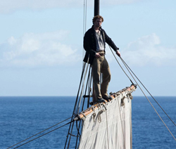 A man in period clothing stands on a wooden yardarm of a sailing vessel overlooking the ocean.