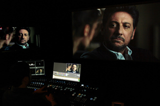 A person works at a control console in a dark room with large screens showing a man's face.