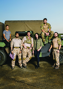 Six people in military and casual attire pose in front of a large green military vehicle outdoors.