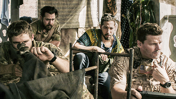 Four men in military and casual clothing look on with concerned expressions in a cluttered outdoor setting.