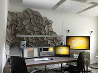 A desk with two computer monitors, a speaker, and a large grey textured wall art piece behind it.