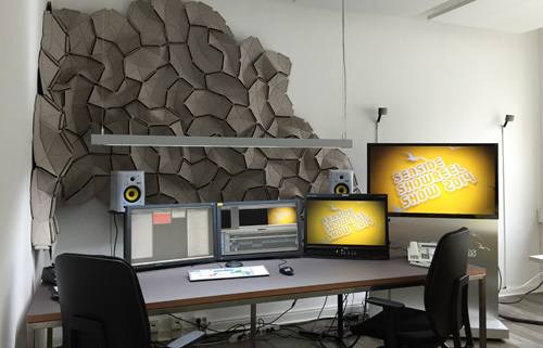 Video editing desk with three monitors, studio speakers, and large geometric acoustic wall panels.