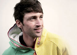 A man with dark hair wearing a yellow and green checkered zip-up jacket looking off-camera.