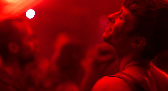 A person with eyes closed and a wide smile, illuminated by intense red lighting in a dark setting.