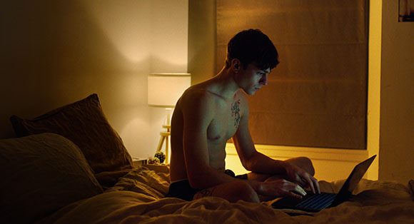 A shirtless young man sits on a bed in a dark room, looking at a laptop screen.
