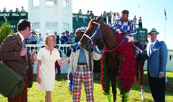 A group of people stand around a brown racehorse draped in red flower garlands on a sunny day.
