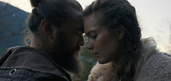 A man with a beard and a woman with braided hair touch foreheads in a close-up profile shot.