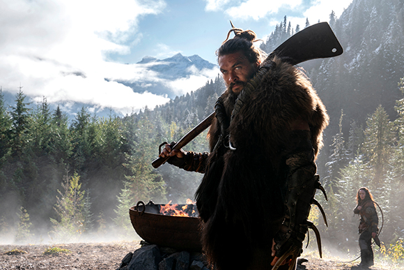 A man with long hair and a fur cloak holds a large primitive blade against a backdrop of mountains and trees.