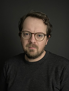 A man with brown hair, glasses, and a beard wearing a dark grey shirt against a black background.