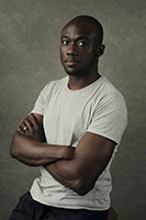 A Black man with a shaved head wearing a white t-shirt, posing with arms crossed against a gray background.