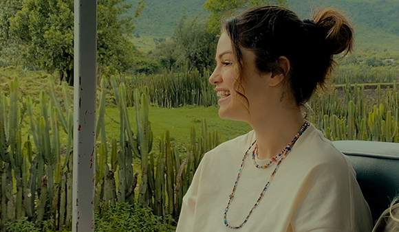 A smiling young woman with dark hair in a bun looks out at a field of tall cacti under a green mountain.
