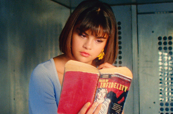 A young woman with a brown bob haircut reads a red book titled 'Tales of Infidelity' in a dimly lit setting.