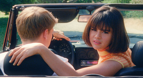 A woman with dark hair looks over her shoulder from a convertible car while a man drives.