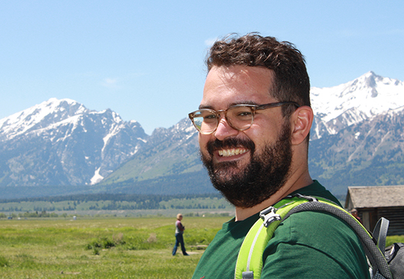 A smiling man with dark hair, a beard, and glasses stands before snow-capped mountains and a green valley.