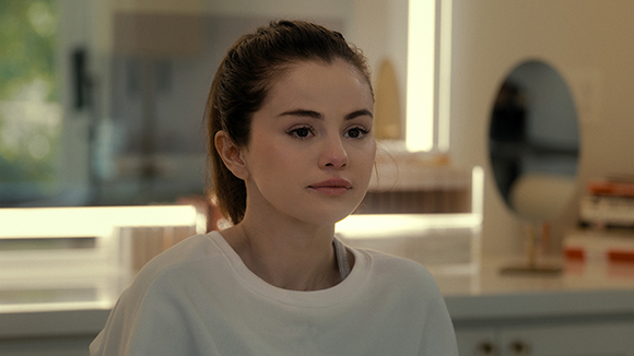 A woman with dark hair pulled back wears a white shirt and looks thoughtfully into the distance.