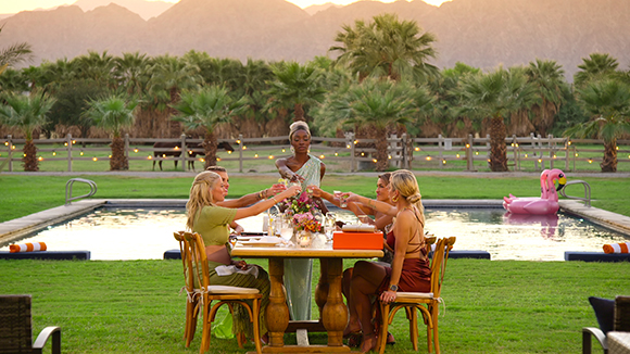 Five women sit around a wooden table dining and drinking by a swimming pool with palm trees and mountains behind.