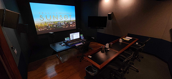 A dark screening room featuring a large projection screen, a mixing console, and a long viewing desk.