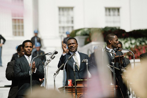A man in a dark suit speaks into microphones at a podium, flanked by men in suits.