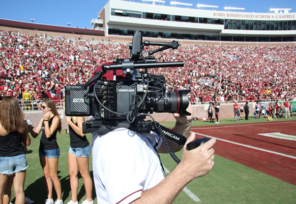 A person operates a large professional video camera overlooking a crowded sports stadium.