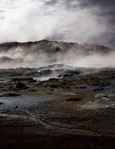 Steam rises from dark, rocky ground in a geothermal field under a cloudy sky.