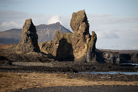 Large, jagged rock formations stand on a dark, rocky coastline under a cloudy sky with mountains behind.