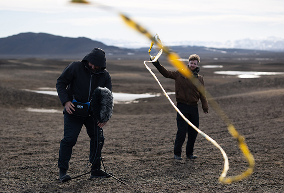 A man adjusts audio equipment while another man holds up a long yellow strip in a wide, open field.
