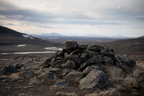 A pile of dark, lichen-covered rocks in the foreground of a wide, barren valley under a cloudy sky.