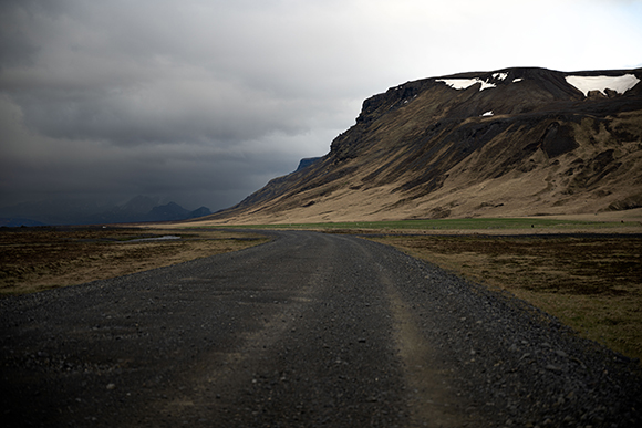 A gravel road curves through a wide, grassy plain toward large, dark mountains under a heavy, overcast sky.