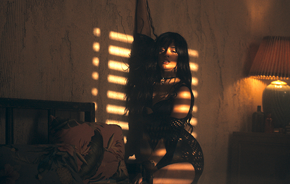 A woman in dark lingerie sits in a dimly lit room with sunlight streaming through blinds.