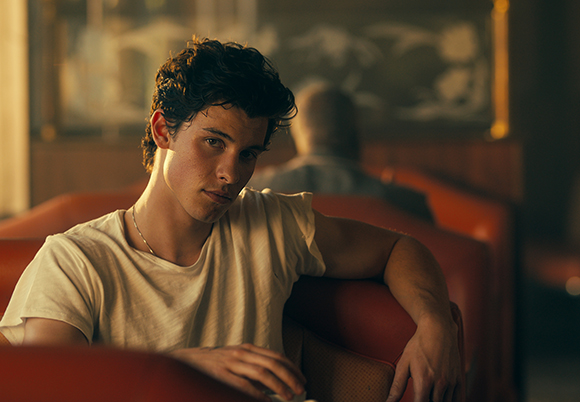 A young man with dark wavy hair wearing a white t-shirt sits in a red booth under warm lighting.