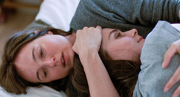 Two women lying close together on a white bed, one resting her head near the other's face.