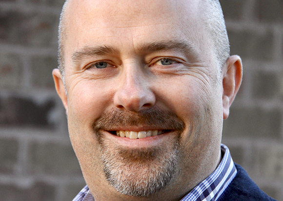 A close-up headshot of a smiling man with light eyes and a salt-and-pepper goatee against a brick background.