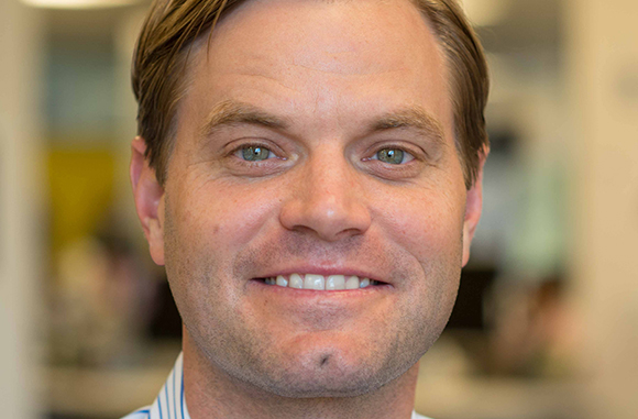 A smiling man with light brown hair and green eyes looking directly at the camera.