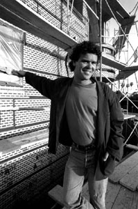 A smiling man in a jacket and t-shirt stands before a brick building with scaffolding.