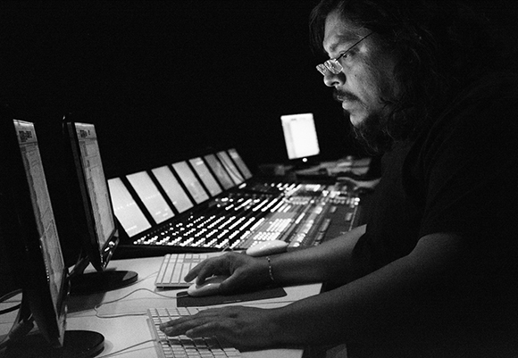 A man with glasses and a beard operates a large mixing desk and computer monitors in a dark room.