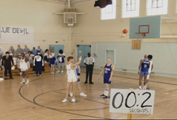 Basketball players stand on a court as a ball flies toward a hoop; a scoreboard shows 00:2 seconds.