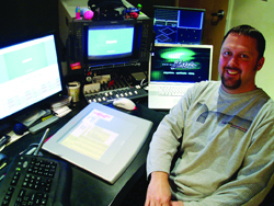 A smiling man sits at a workstation equipped with multiple monitors, a graphics tablet, and editing controls.