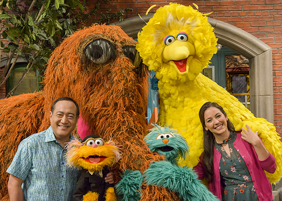 A man and woman smile while posing with large orange, yellow, and teal furry Sesame Street characters.