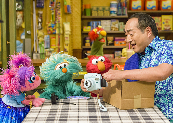 A man in a blue floral shirt opens a box with a light blue instant camera at a table with colorful puppets.