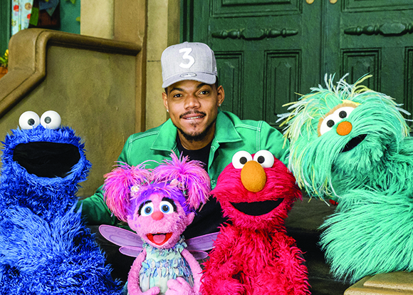 A man in a green shirt and grey cap sits between Cookie Monster, Abby Cadabby, Elmo, and Rosita.
