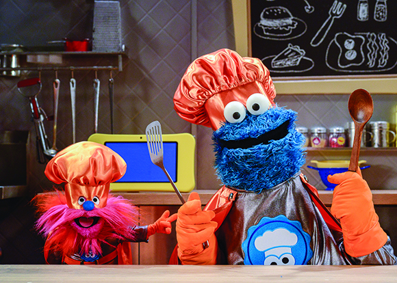 A large blue fuzzy puppet and a small pink fuzzy puppet wearing orange chef hats and holding kitchen utensils.
