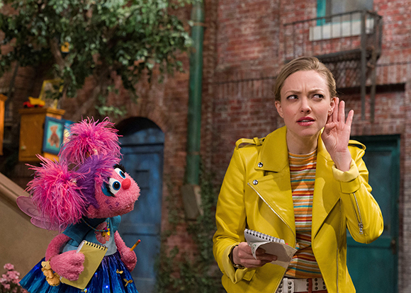 A woman in a yellow jacket looks startled at a pink fuzzy puppet in a courtyard.