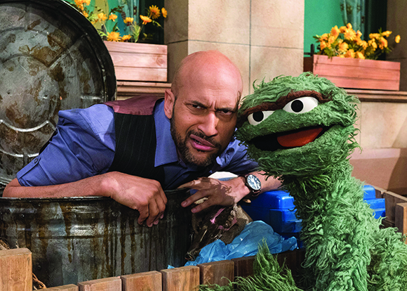 A bald man with a beard and a green fuzzy puppet peek out from behind a large metal trash can.