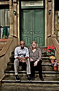 A man in a light blue shirt and a woman in a patterned blouse sit on stone steps in front of a green door.