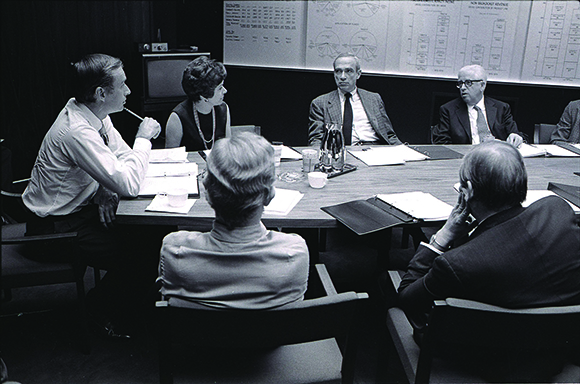 A group of people in business attire sit around a large table in a meeting room with charts on the wall.