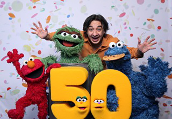 A man smiles with Elmo, Cookie Monster, and Oscar the Grouch in front of a confetti background and a large number 50.