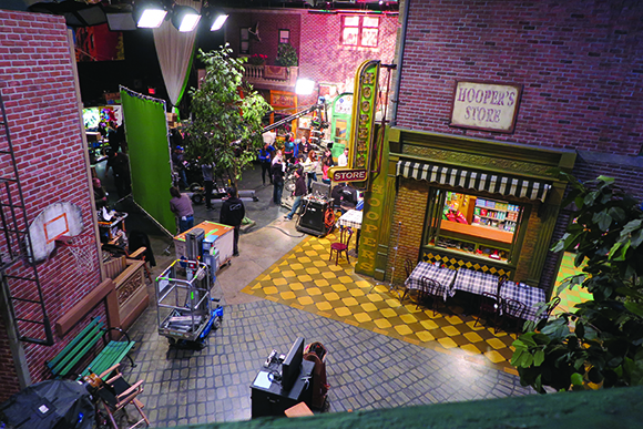 High-angle view of a TV studio set with a brick storefront, street props, and crew members working.