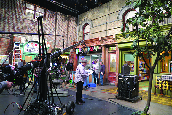 Film crew and camera equipment on a studio set designed to look like a city street with storefronts.