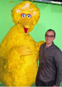 A man in a grey shirt smiles while standing next to the large yellow character Big Bird against a green screen.