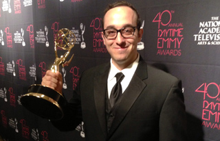 A man in a black tuxedo and glasses smiles while holding a gold Emmy Award trophy.